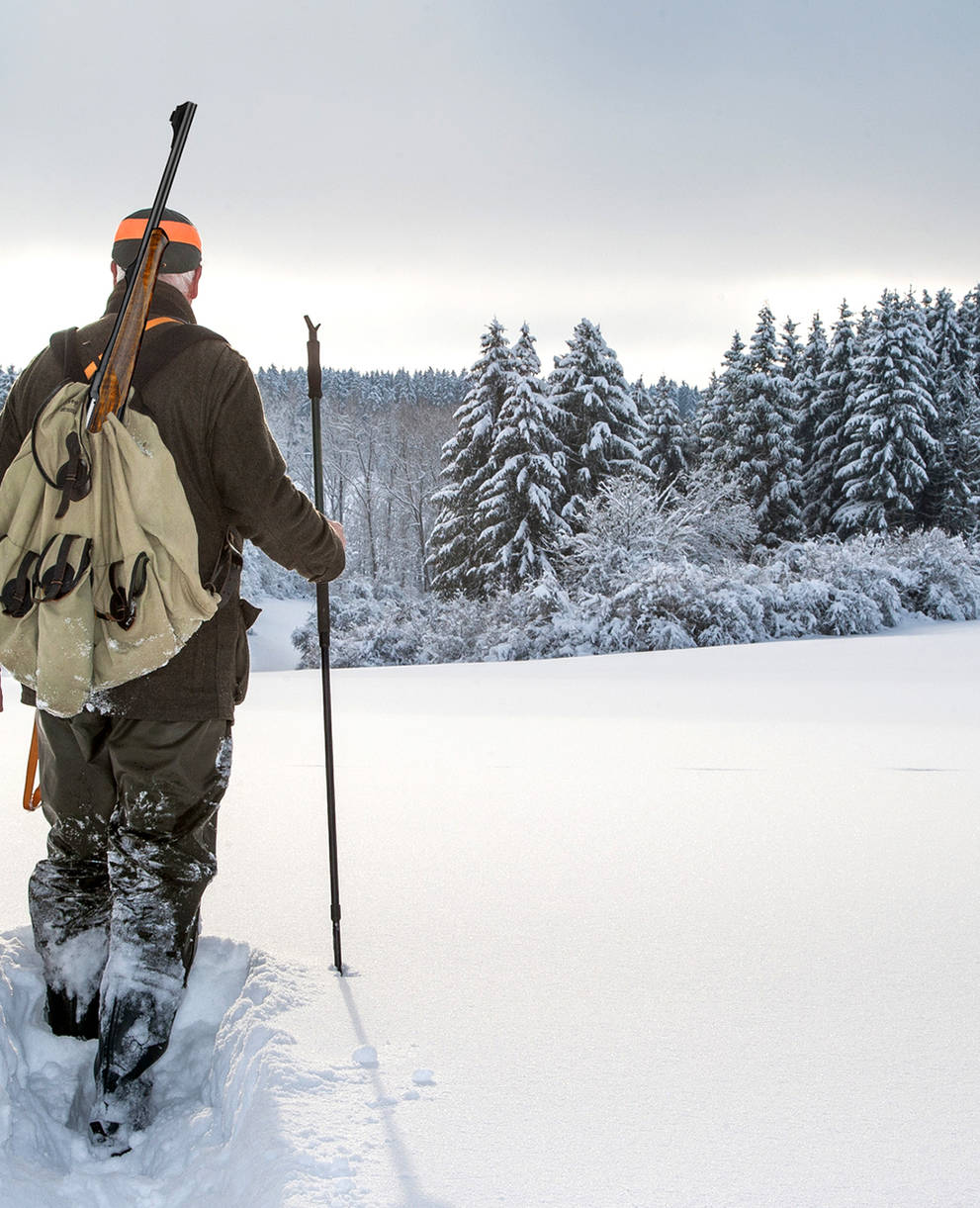 In einer Winterlandschaft geht ein Jäger durch den frischen Schnee hinunter ins Tal.